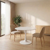 White tulip dining table with two beige upholstered chairs angled around it in a sunlit room