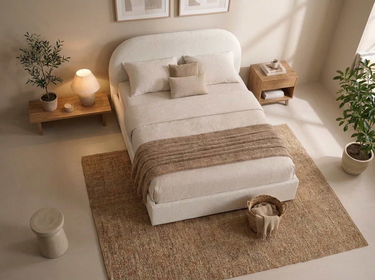 A modern upholstered bed with a rounded headboard, dressed in cream bedding and a taupe throw blanket, styled in a warm, minimalist bedroom with a woven rug, wooden side tables, and potted plants.