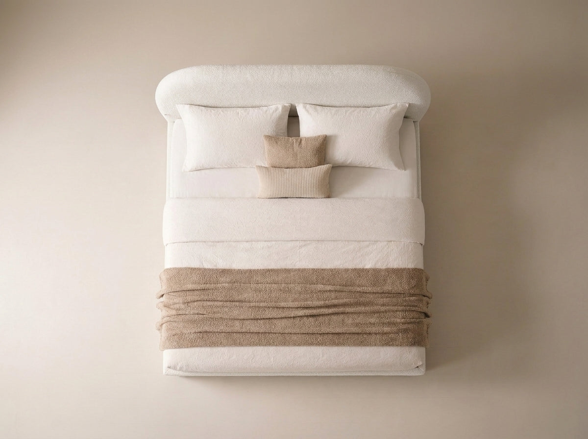 A top-down view of a neatly made bed with a white upholstered headboard, white bedding, two white pillows, two beige accent pillows, and a beige throw blanket wrapped around the foot of the bed against a warm beige wall.