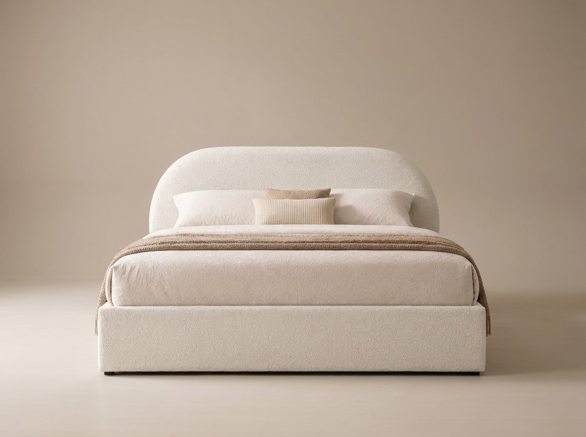 A modern platform bed with a rounded headboard and upholstered base, dressed in cream bedding and a beige throw blanket, set against a minimalist beige background.